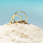Pair of gold wedding ring bands jewelry in sand on tropical desert island beach during summer with blue ocean background
