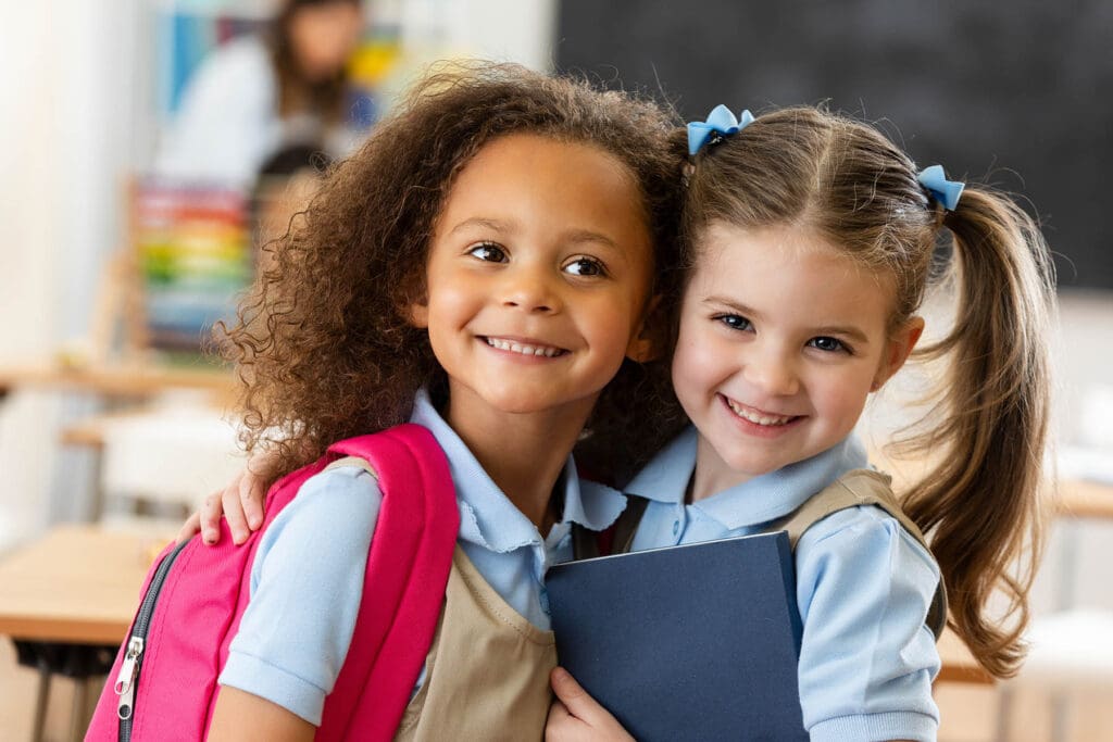 Two smiling girls in school uniforms, carrying backpacks and books, embody friendship and joy in a classroom setting.