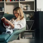 A smiling Caucasian female resting in her living room while enjoying reading a book.